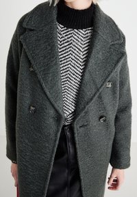 Abrigo de doble botonadura en verde oscuro, hecho de tela texturizada, superpuesto a un suéter de patrón en blanco y negro, combinado con pantalones de cuero negros.