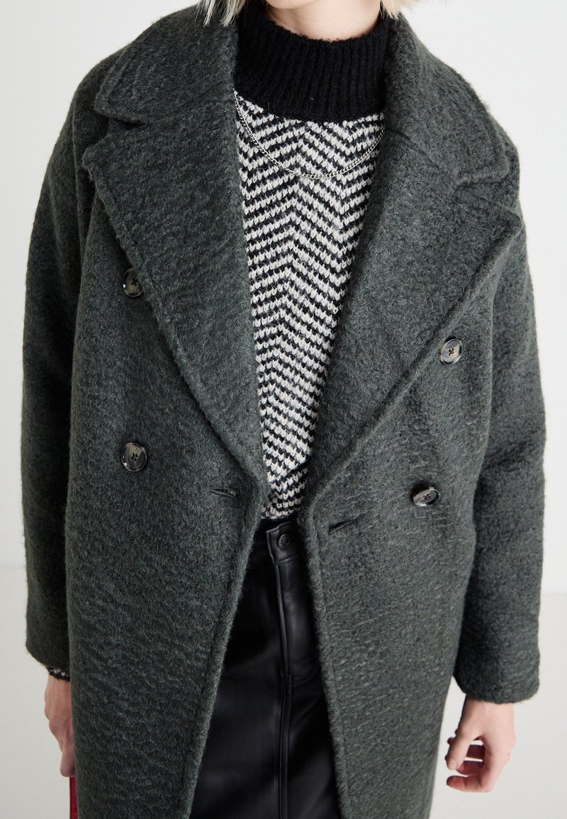 Abrigo de doble botonadura en verde oscuro, hecho de tela texturizada, superpuesto a un suéter de patrón en blanco y negro, combinado con pantalones de cuero negros.