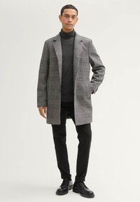 Grau karierter Übermantel mit Reverskragen, getragen über einem dunklen Rollkragenpullover, kombiniert mit schwarzen Hosen und schwarzen Schnürschuhen. Schlankes Design.