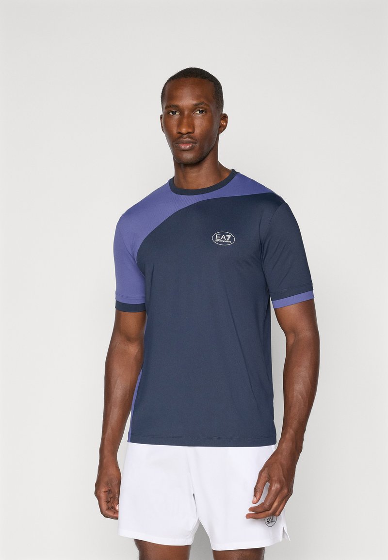 T-shirt atlético azul marinho com detalhes roxos, gola redonda e mangas curtas. Apresenta o logótipo EA7 no peito e um tecido texturizado.