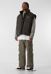 Schwarwest, puffy, ärmellose Weste mit diagonalen Nähten und hohem Kragen. Getragen über einem grauen Pullover, einem weißen Hemd und olivfarbenen Cargo-Hosen.