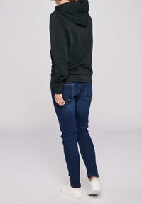 Svart huvtröja med dragsko, kombinerad med mörkblå slim fit-jeans. Klädseln avslutas med vita sneakers. Texturen ser mjuk ut.