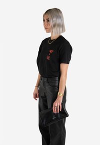 Schwarzes grafisches T-Shirt mit roten Drucken, kombiniert mit schwarzen Lederhosen. Hält eine schwarze Clutch-Tasche in der Hand, goldene Uhr am Handgelenk. Kurze, helle Haare.