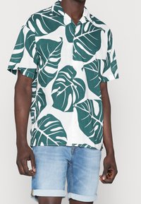 Camicia a maniche corte con bottoni di colore bianco, decorata con un grande motivo di foglie verdi, realizzata in tessuto leggero, abbinata a pantaloni shorts in denim arrotolati.