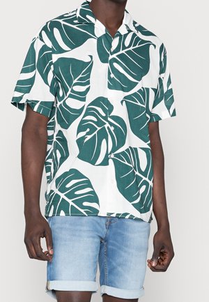 Chemise à manches courtes boutonnée blanche avec un grand motif de feuilles vertes, en tissu léger, associée à un short en denim retroussé.