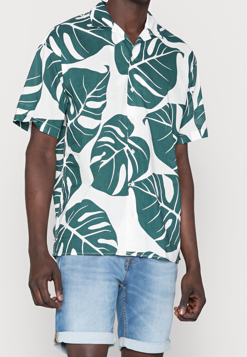 Camicia a maniche corte con bottoni di colore bianco, decorata con un grande motivo di foglie verdi, realizzata in tessuto leggero, abbinata a pantaloni shorts in denim arrotolati.