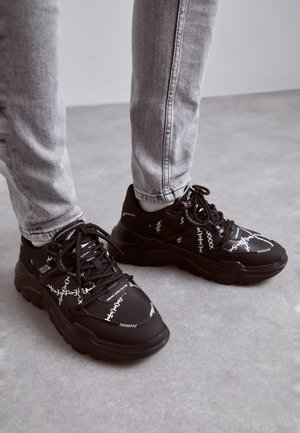 Person trägt graue Jeans und schwarze klobige Sneaker mit weißem Aufdruck und steht auf grauem Boden.