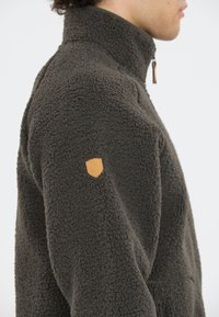 Fleecejacka i mörkgrön med hög krage, texturerat pälsliknande material och en liten brun läderlogga på ärmen.