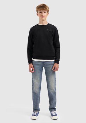 Jonge man staat, gekleed in een zwart sweatshirt, lichtblauwe jeans, wit T-shirt en blauwe sneakers tegen een effen grijze achtergrond.