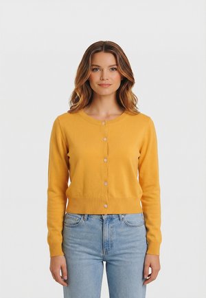 Donna con capelli castani ondulati che indossa un cardigan giallo senape con bottoni e jeans azzurri, in piedi davanti a uno sfondo bianco uniforme.