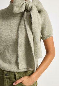 Ljusgrön stickad kortärmad tröja med hög krage och en stor rosett knuten vid halsen, med en mjuk, fuzzy textur.