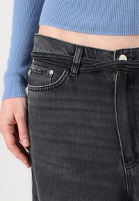 Black denim jeans with a high waist, featuring a classic five-pocket design, metal hardware, and a stitched waistband. Blue ribbed sweater visible.