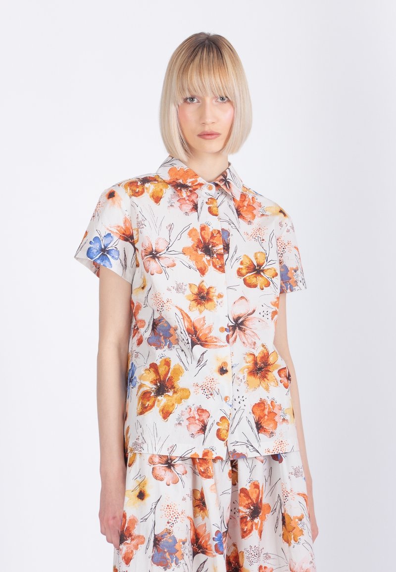 Joven con bob rubio lacio que lleva una camisa blanca de manga corta con botones y una falda con estampado floral en naranja, amarillo y azul.