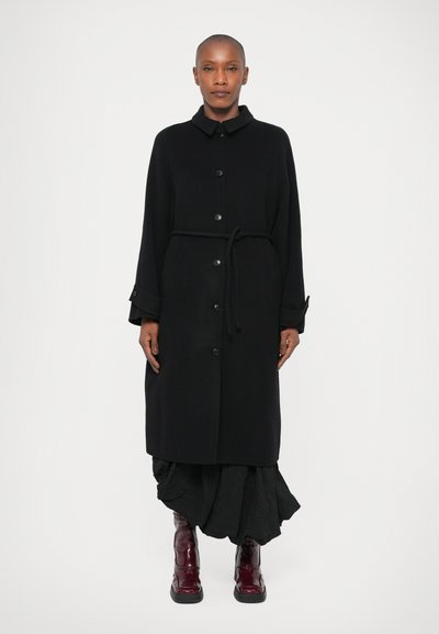 Schwarzer Mantel mit Kragen, Knöpfen auf der Vorderseite und einem Gürtel in der Taille. Das Modell trägt einen mehrlagigen Rock und glänzende weinrote Stiefel.
