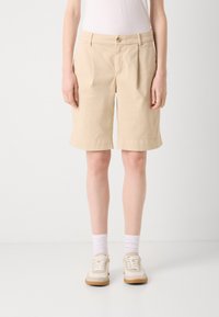 Pantaloni corti di cotone beige con un taglio dritto, dotati di tasche anteriori, chiusura con bottone e orli arrotolati. Indossati con calzini bianchi e sneaker.