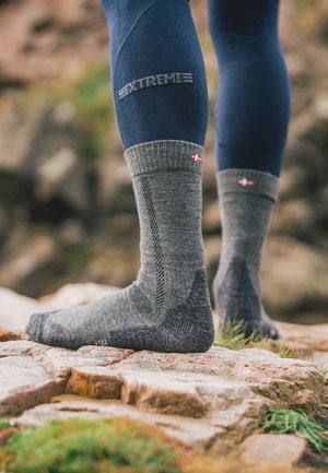 Graue Wollsocken mit verstärktem Fersen- und Zehenbereich, geripptem Bündchen und einem kleinen Flaggenakzent an der Oberseite, die auf felsigem Terrain liegen.