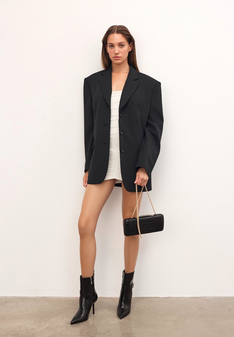 Blazer nero oversize con spalle strutturate, abbinato a un vestito bianco aderente. Accessoriato con una borsa nera con effetto coccodrillo e stivali con tacco.