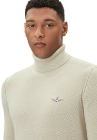 Maglione beige a collo alto in maglia con motivo testurizzato e un piccolo ricamo di un emblema di aquila sul petto sinistro.