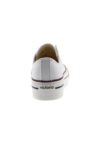 Zapatilla deslizante de lona blanca con suela plana de goma, que presenta una franja marrón y la marca "victoria" en el talón. Textura suave.