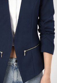 Námořnický blazer s strukturovaným designem, dvěma zipovými kapsami se stříbrným kováním a ohrnutými rukávy přes bílou košili s knoflíky.