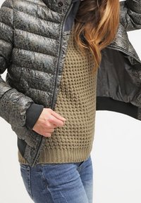 Kurtka pikowana w ciemnozielonym kolorze z teksturowanym wzorem w panterkę; zestawiona z grubym, oliwkowozielonym swetrem z dzianiny i niebieskimi dżinsami.