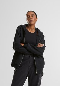 Felpa nera con zip, dal tessuto morbido, con zip frontale, tasche a marsupio e vestibilità comoda, abbinata a una canotta nera e jeans.