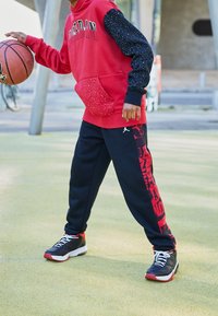 Červená mikina s kapucí a černými rukávy s tečkami, vybavená přední kapsou. Černé tepláky s červeným logem. Černé a červené tenisky, držící basketbal.