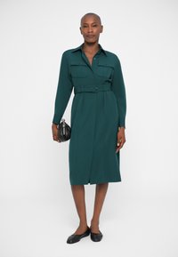 Tume roheline pluusikleit pika varrukaga, kahe rinnatasku ja vöödiga talje, põlvini ulatuva seelikuosa. Venitatud mustade lamedate kingade ja väikese käekotiga.