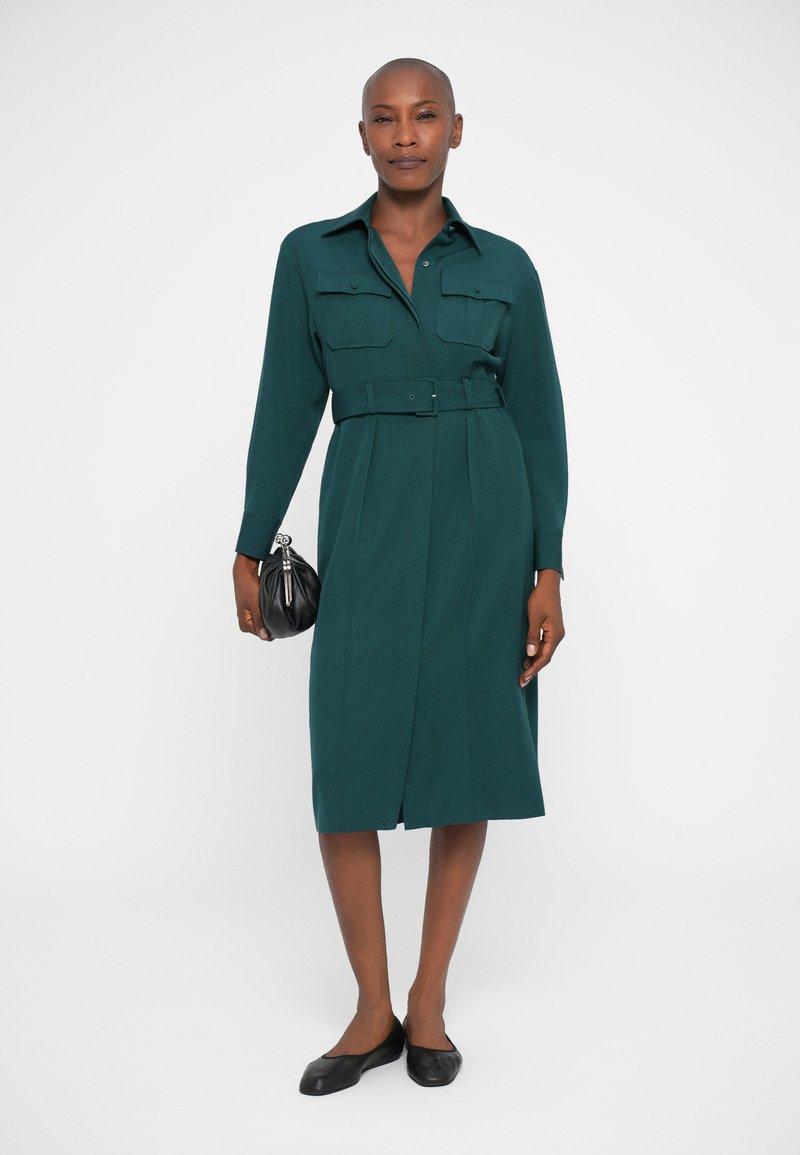 Tume roheline pluusikleit pika varrukaga, kahe rinnatasku ja vöödiga talje, põlvini ulatuva seelikuosa. Venitatud mustade lamedate kingade ja väikese käekotiga.