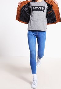 Osoba w niebieskich wąskich dżinsach, białych trampkach, szarym podkoszulku Levi's i rozpiętej brązowej kurtce, idąca do przodu na jasnym tle.