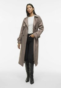 Lungo trench coat marrone chiaro con vita sbloccata e spalline sulle spalle, abbinato a pantaloni neri aderenti e stivali neri alti fino al ginocchio.