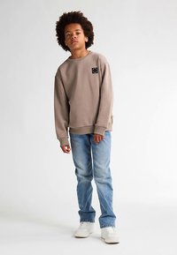 Felpa marrone chiaro con scollo tondo e una piccola toppa con logo, abbinata a jeans azzurri e scarpe da ginnastica bianche. Tessuto liscio, vestibilità casual.