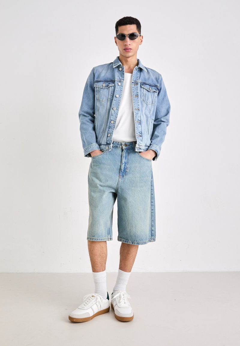 Giacca di jeans e pantaloni corti di un blu chiaro, con tasche e una vestibilità rilassata. Abbinati a sneakers bianche e occhiali da sole.