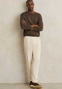 Brauner langer Pullover, beigefarbene, plissierte Hose und braune Schuhe. Das Modell steht mit verschränkten Armen vor einem neutralen Hintergrund.