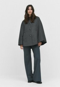 Manteau en mélange de laine grise avec un col montant, comportant cinq boutons noirs et des poches latérales, associé à un pantalon gris sur mesure.
