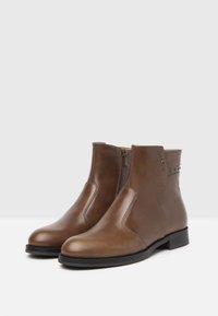 Stivaletti ankle in pelle marrone con finitura liscia, zip laterale, borchie decorative e tacco basso a blocco. Design con punta arrotondata chiusa.