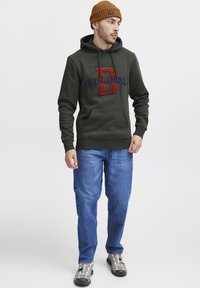 Green hoodie with “D ORIGINAL” embroidered in red and blue, front pocket, paired with blue jeans and gray sneakers, styled with a brown beanie.
