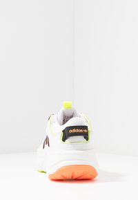 Vit Adidas-sneaker med orange sula och logotyp på hälfliken, neongrän accentuering, visad bakifrån på en vit yta mot en vit vägg.