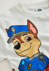 Chien policier de dessin animé souriant portant un uniforme bleu et un chapeau avec un badge en forme d’empreinte de patte, imprimé sur un vêtement en tissu blanc.