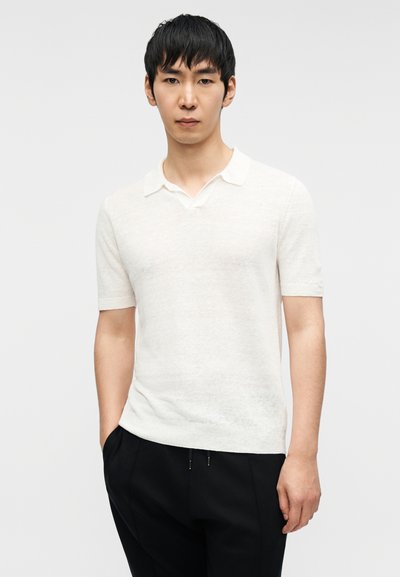 Άσπρο polo με κοντά μανίκια και V-λαιμόκοψη με ριμπ ύφανση στο γιακά, κατασκευασμένο από υφασμα με υφή. Φοριέται με μαύρο παντελόνι, το μοντέλο στέκεται όρθιο.