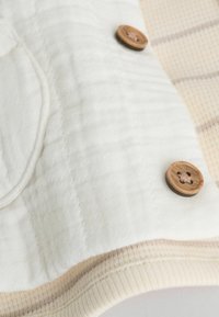Tela blanca acolchada con una textura suave, que presenta un botón de madera y un acento rayado beige en el borde.