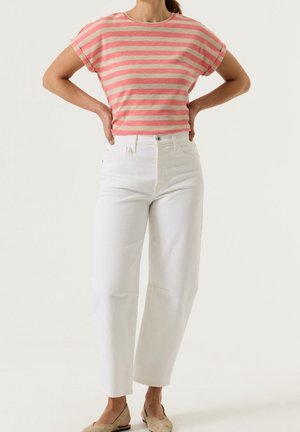 Femme portant un haut rayé rose et beige à manches courtes, un pantalon blanc taille haute et des chaussures plates beiges, debout les mains sur les hanches.