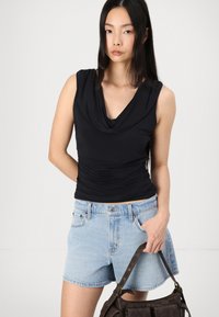 Zwarte mouwloze top met een colhals en geplooide textuur, gecombineerd met lichtblauwe spijkerbroekshorts. Houdt een donkerbruine handtas vast.