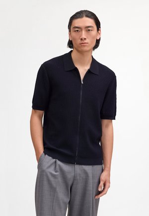 Unge mand iført sort tekstureret kortærmet skjorte med lynlås og grå plisserede bukser, stående med den ene hånd i lommen mod en ensfarvet baggrund.