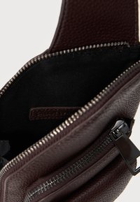 Σκουρό καφέ δερμάτινη τσάντα με υφή, με μερικώς ανοιχτό φερμουάρ που αποκαλύπτει εσωτερική επένδυση από μαύρο ύφασμα και έναν δερμάτινο ιμάντα στο εσωτερικό.