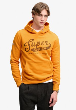 Ung mand med lysebrunt hår iført en orange hættetrøje med teksten "Super Dry 1954" og mørkegrå bukser, stående med den ene hånd i lommen.