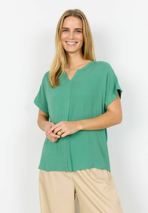 Smilende kvinde med langt blondt hår iført en kortærmet grøn bluse og beige bukser, stående med hænderne foldet.