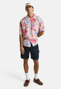 Korte mouwen shirt met een multicolore tropische print in roze en blauw, gecombineerd met donkere cargoshorts, witte sokken en bruine schoenen.