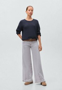 Pulover din tricot navy cu textură canelate, blugi gri deschis with picior larg, curea maro cu cataramă rotundă și pantofi bej. Fundal simplu.