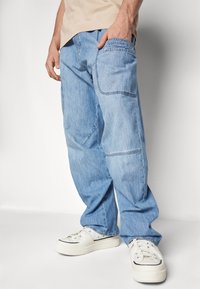 Svetlo modri jeans s sproščenim fitom, z žepom na strani in diskretnimi šivi. V kombinaciji z belimi supergami.